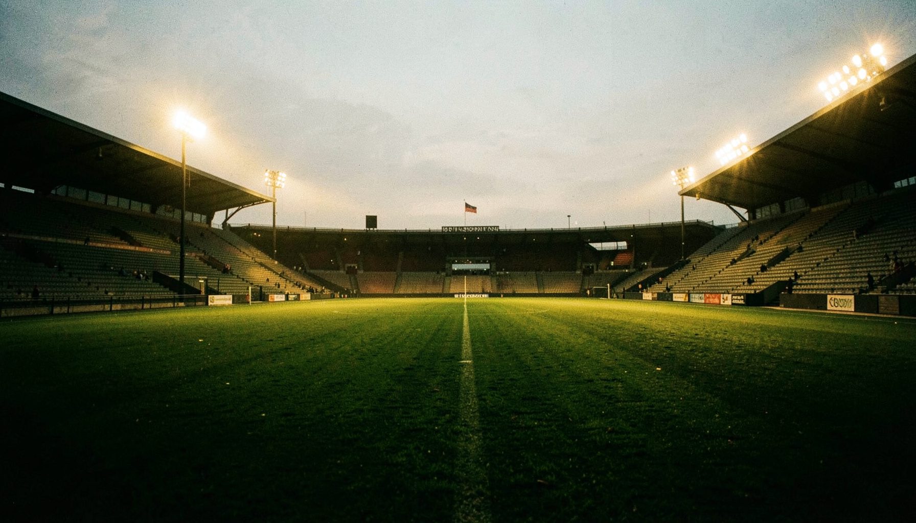Wide documentary photograph of a floodlit football pitch at dusk, deep green turf under warm amber stadium lights, empty stands in shadow, moody and atmospheric.