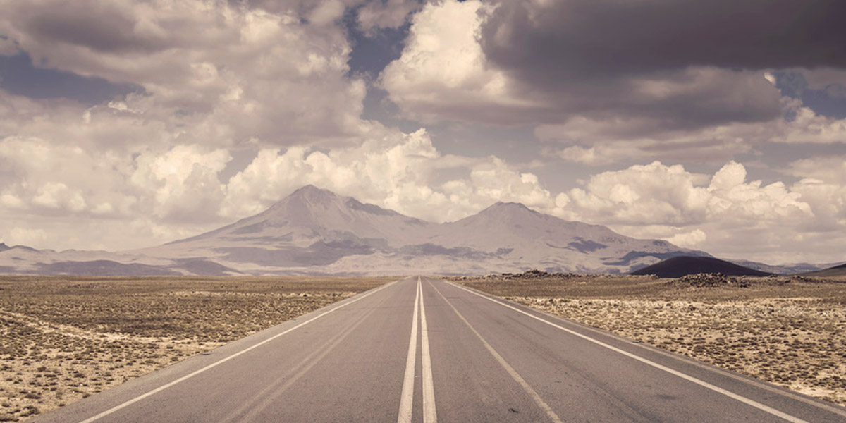 Empty paved road stretching toward distant mountains under a cloudy sky, muted sepia tones, calm and vast mood